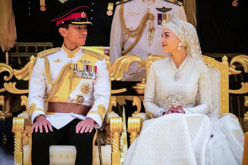 Royal couple at their wedding looking at each other sitting on gold thrones. 