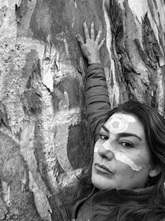 A woman with paint on her face touches a tree. 