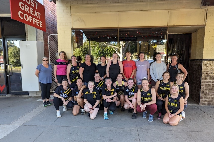 A team of netball players stands out the front of a coffee shop.
