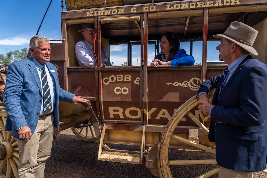A man and a woman sit inside an old-style horse-drawn coach. Two men stand outside leaning on it, chatting.