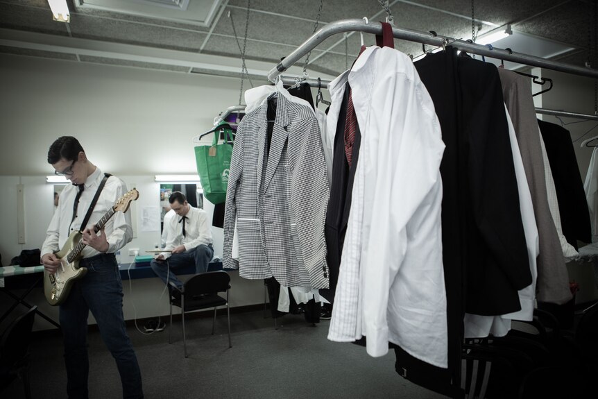 Actors in dressing room practising with guitar and drumsticks, costumes on rack