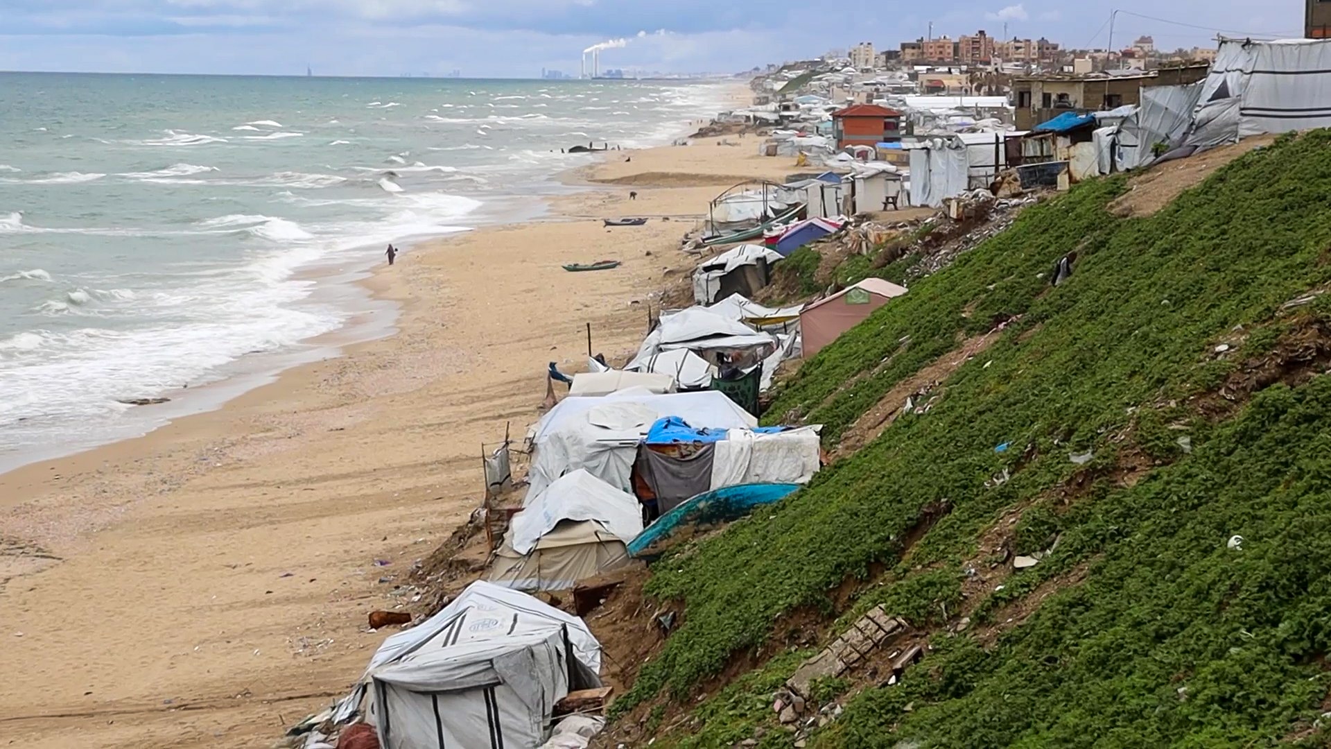 dozens of make-shift tents on the edge of a beach in front of a hill