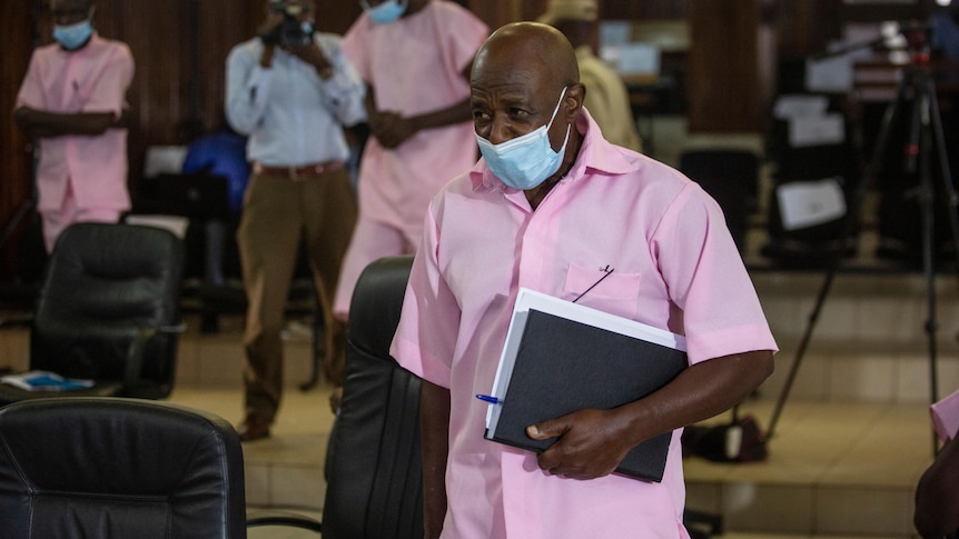 A bald man in a pink short sleeve shirt wears a face mask as he carries a bundle of docments through a courtroom. 
