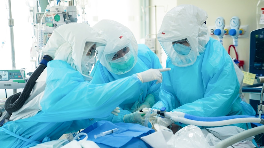 Three doctors and nurses wear protective equipment as they treat a patient in a hospital room.