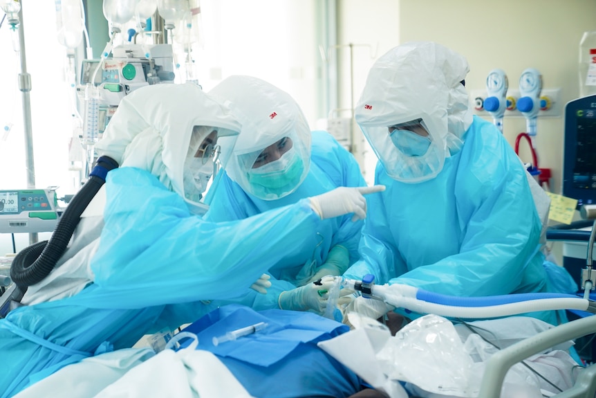 Three doctors and nurses wear protective equipment as they treat a patient in a hospital room.