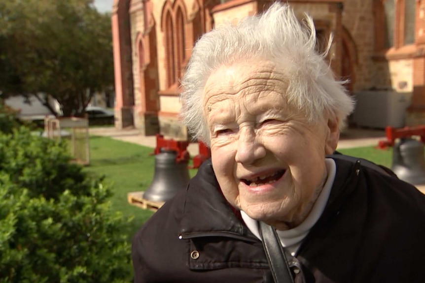 An elderly woman with white hair, with church bells on the grass and a church in the background behind her.