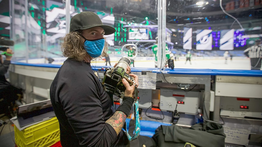 Dave Sandford stands rink side holding a camera and wearing a mask with a game happening behind him.