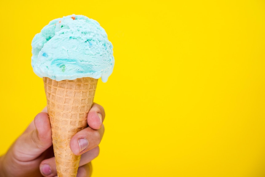A hand holds an ice-cream cone containing blue ice cream. Yellow background. 