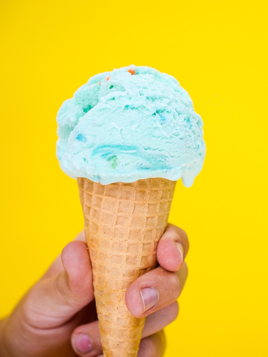 A hand holds an ice cream cone containing blue ice cream. Yellow background. 