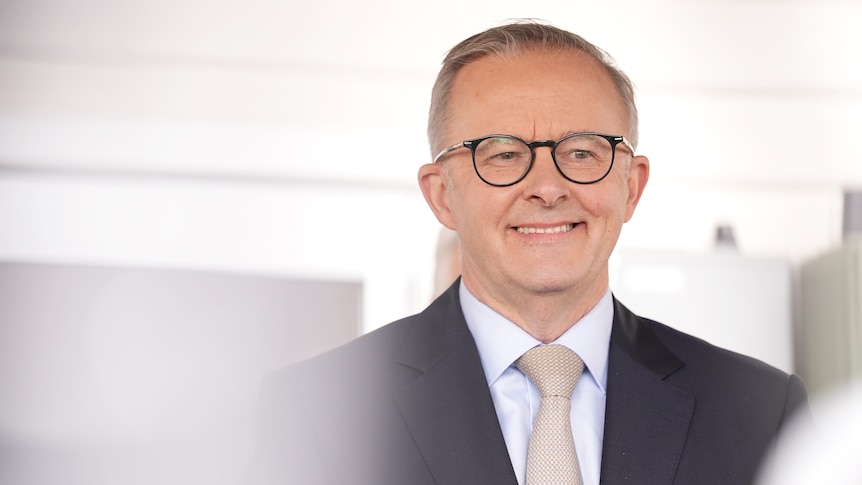 Politician Anthony Albanese stands and smiles, wearing a dark suit and glasses.