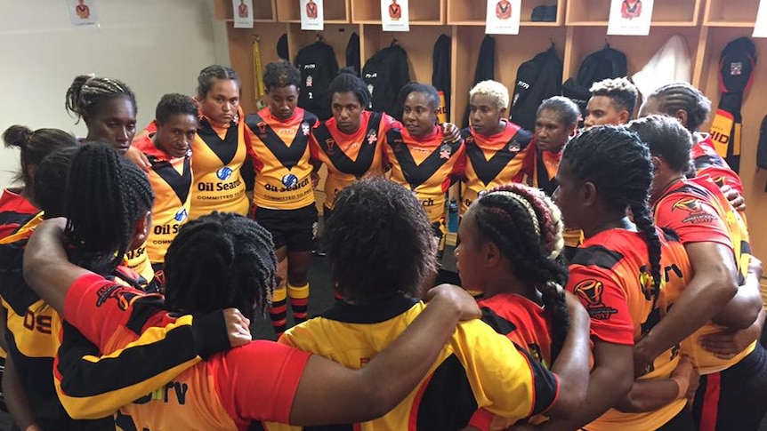 Eighteen women in rugby uniform form a huddle in the middle of the changerooms.