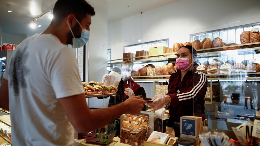 Dark haired woman wearing mask take payment at a shop from tall man wearing mask