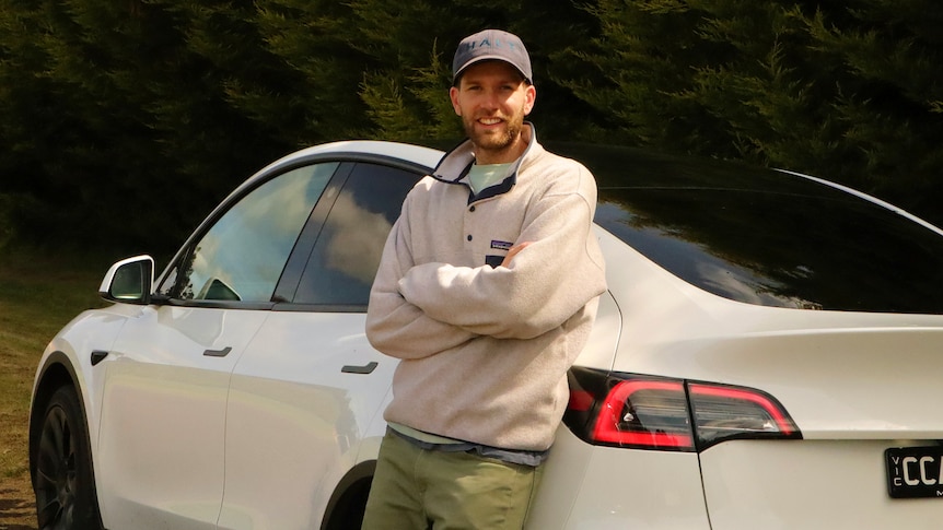A young man leans against a white sedan electric vehicle with arms crossed