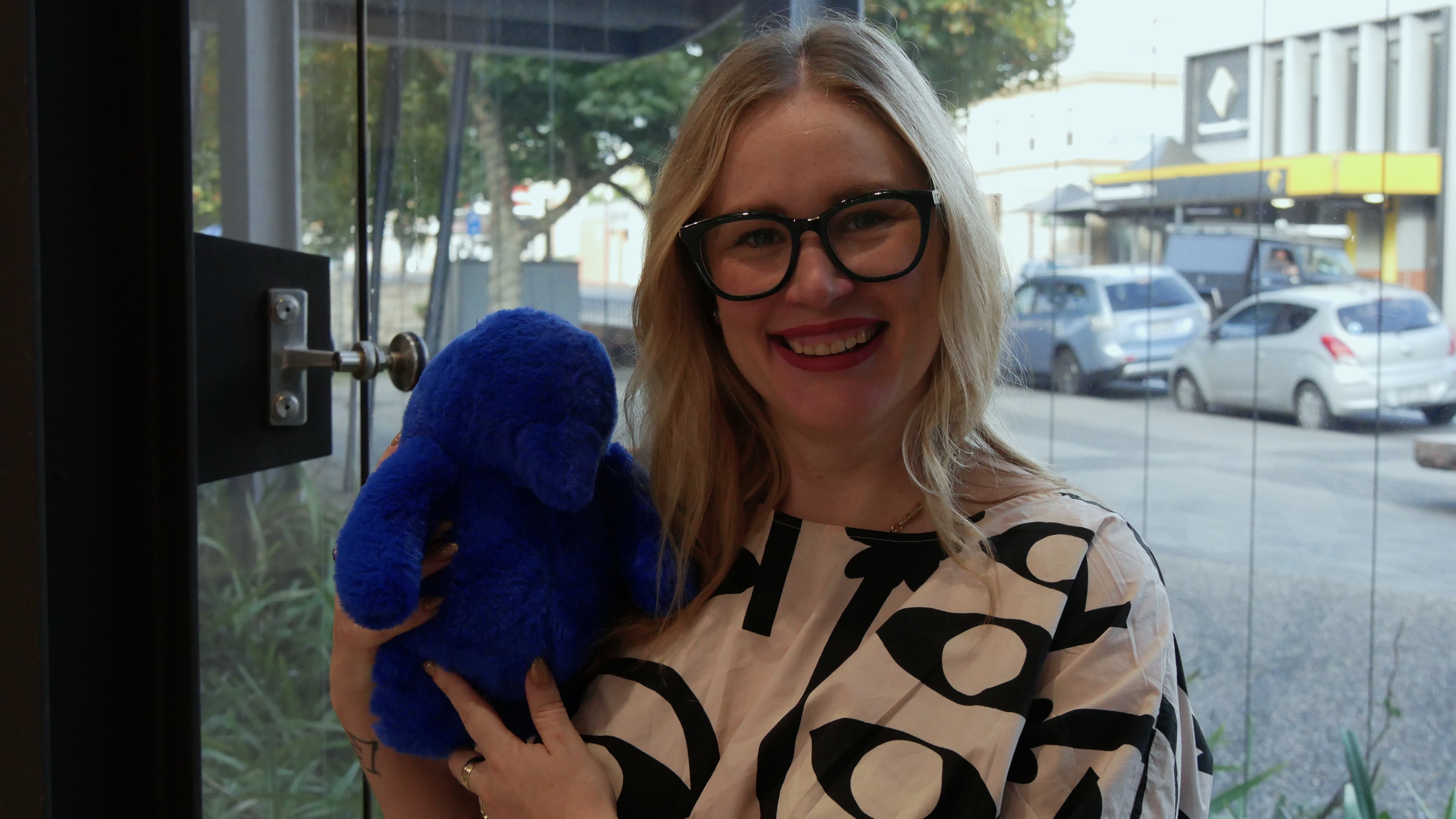 Μια γυναίκα που φορά ένα ασπρόμαυρο τοπ και γυαλιά που κρατά ένα λούτρινο μπλε παιχνίδι.