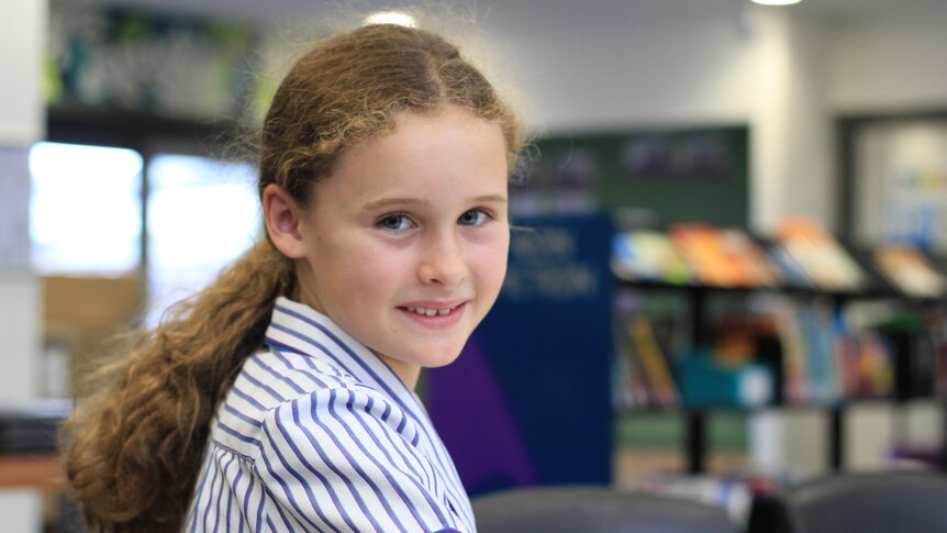 A nine-year-old girl with long fair hair in a striped school uniform in a school library. Smiling.