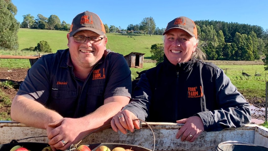 A man and woman wearing blue and orange caps and shirts stand behind a ute tray containing buckets of apples.