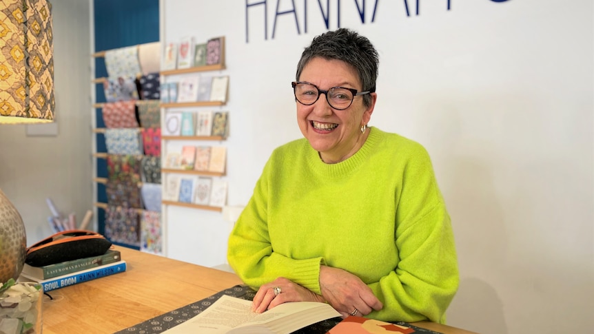 A woman with short dark hair and a bright green jumper smiles at the camera, she stands at a counter with an open book.