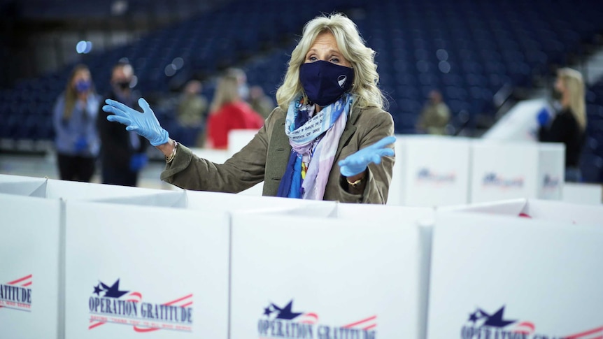 A blonde woman wearing a mask and blue gloves gestures from behind large boxes labelled 'Operation Gratitude'.