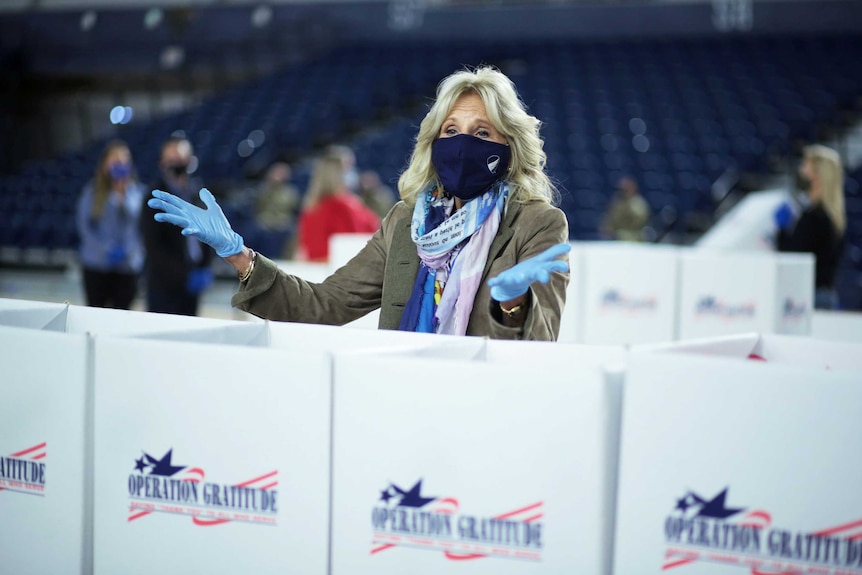 A blonde woman wearing a mask and blue gloves gestures from behind large boxes labelled 'Operation Gratitude'.