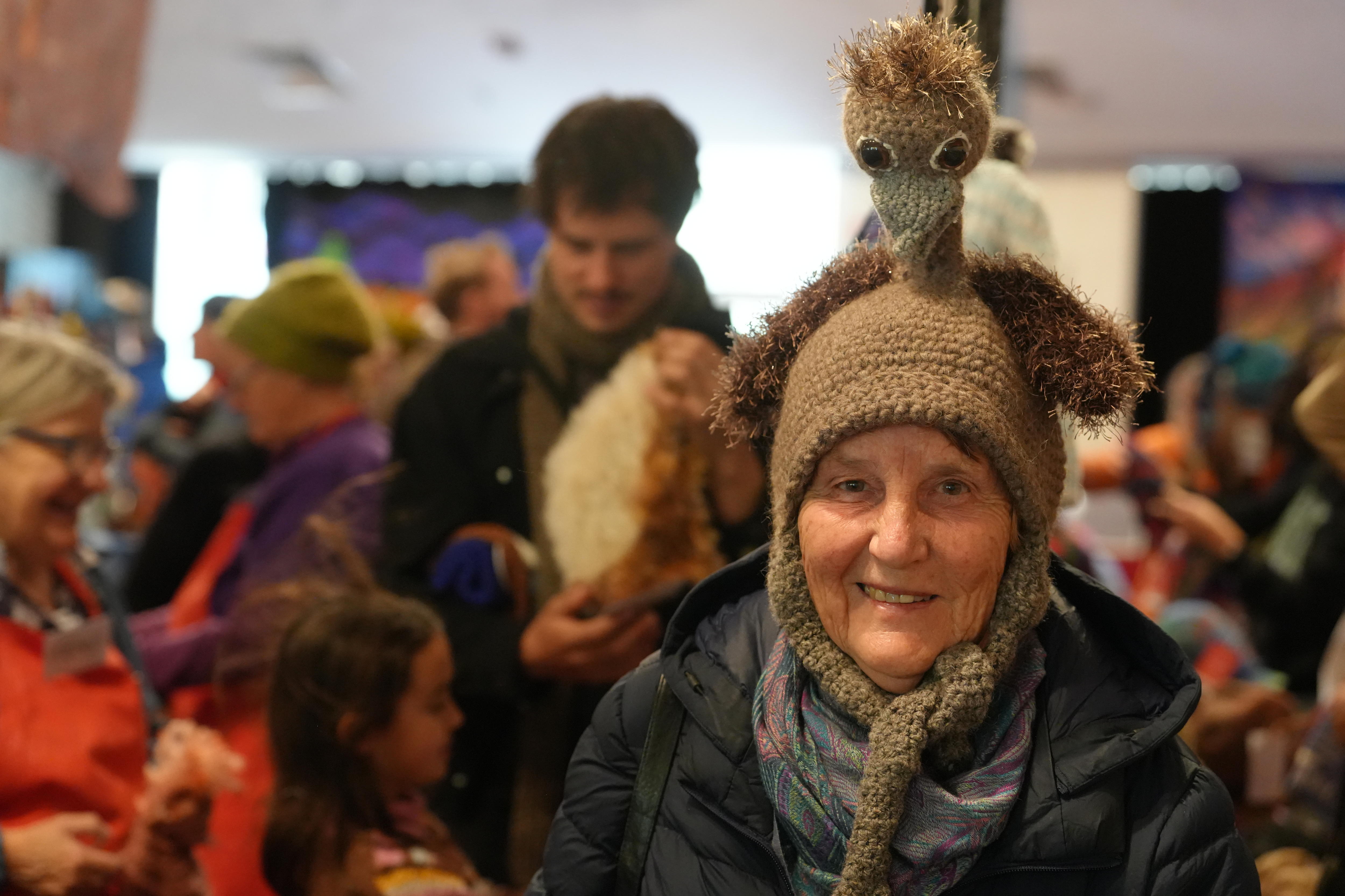 An older woman looking at the camera, wearing a brown beanie with a big emu character knitted to its top.