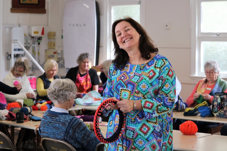 A woman smiles while holding a crochet loom and hook in front of tables of ladies knitting.