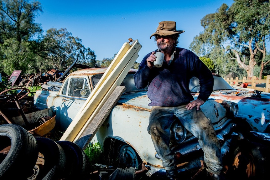 A man sits on a rusty car in a backyard full of wrecks.