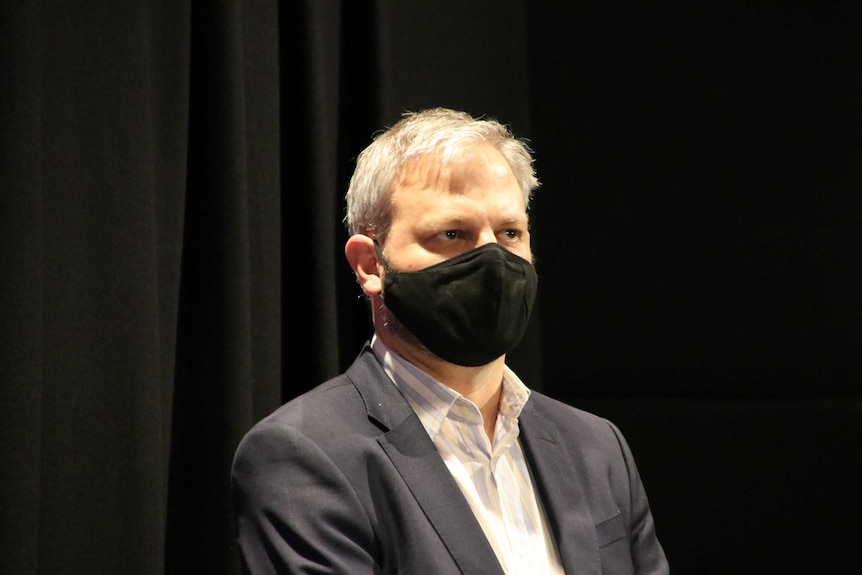 Brett Sutton stands with his hands clasped wearing a black mask and staring ahead.