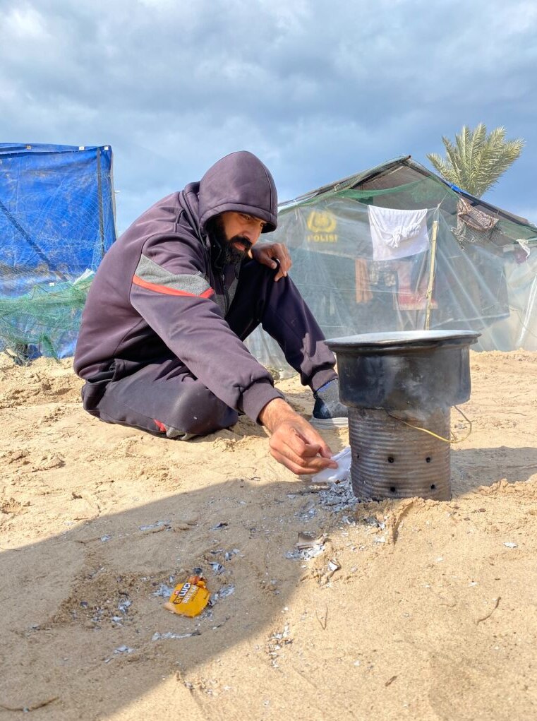 A man sitting in the sand with a tent behind him and small cooking setup in front of him. 