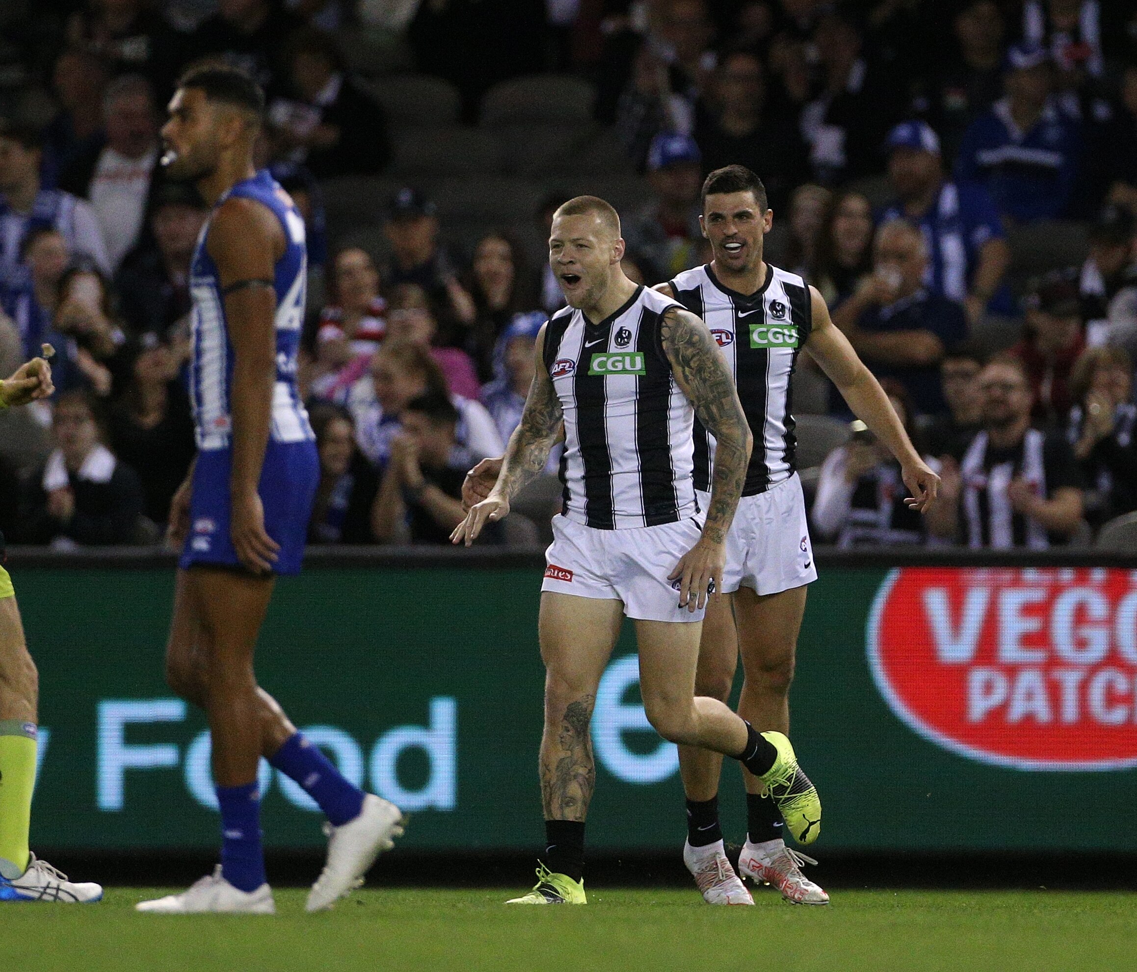 An AFL forward runs back after kicking a goal, with a teammate just behind him celebrating.