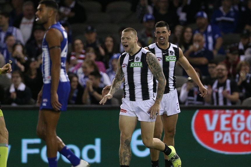 An AFL forward runs back after kicking a goal, with a teammate just behind him celebrating.