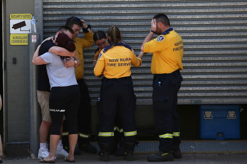 A group of half a dozen people, some of them RFS volunteers, hug and comfort one another outside a shed.