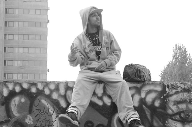 A man wearing a hoodie sits on a graffitied wall.