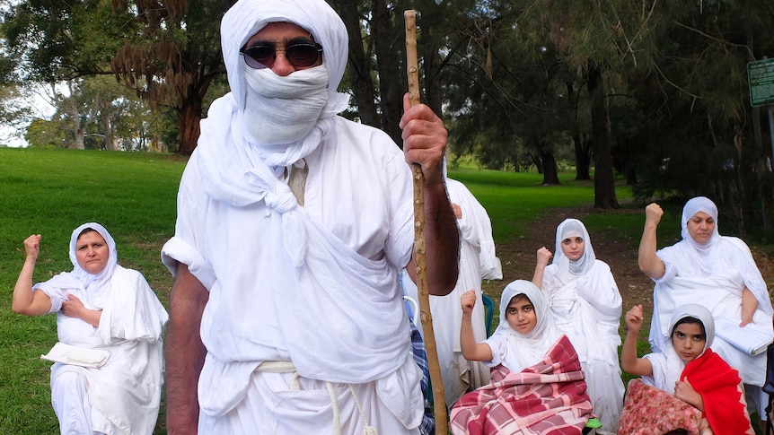 White robed Mandaean leader Rishema Satar Jabar Helo stands with a staff in front of a group of women in robes and blankets.