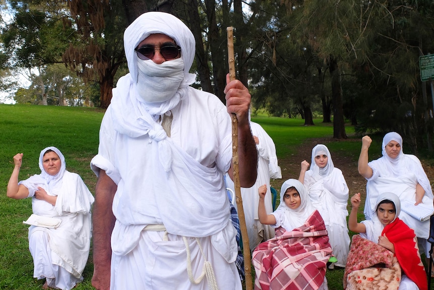 White robed Mandaean leader Rishema Satar Jabar Helo stands with a staff in front of a group of women in robes and blankets.