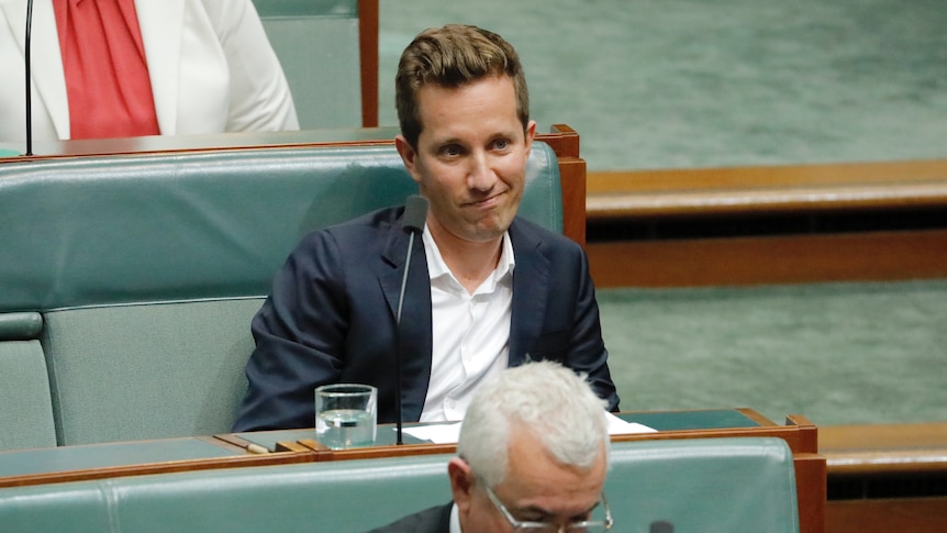 A young man in a suit with no tie grimaces as he sits, listening, in the house of representatives