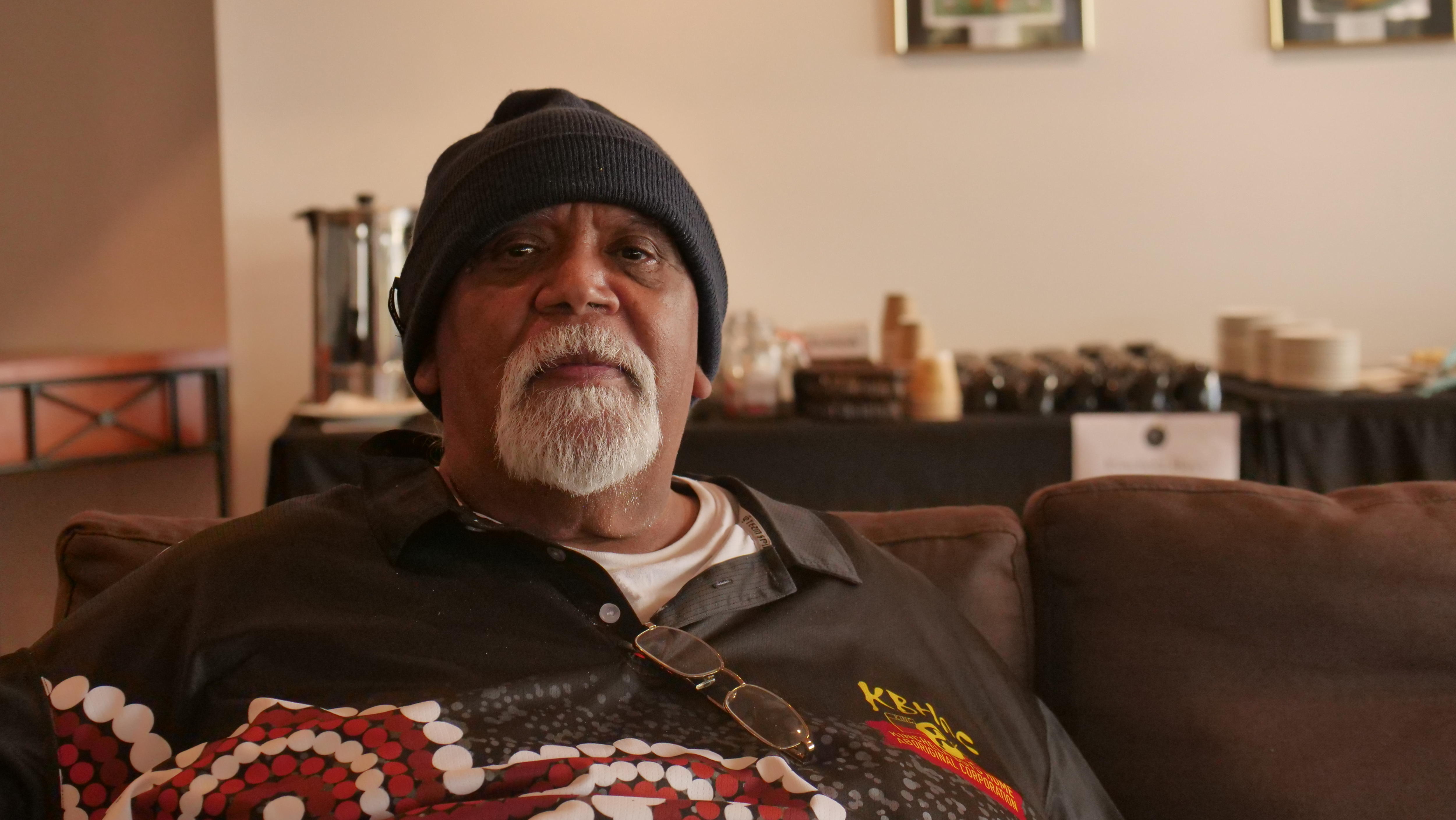 Older man staring solemnly with a black beanie, gray goatie and black, red and white colored shirt