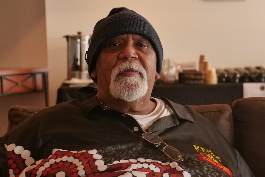 Older man staring solemnly with a black beanie, gray goatie and black, red and white colored shirt