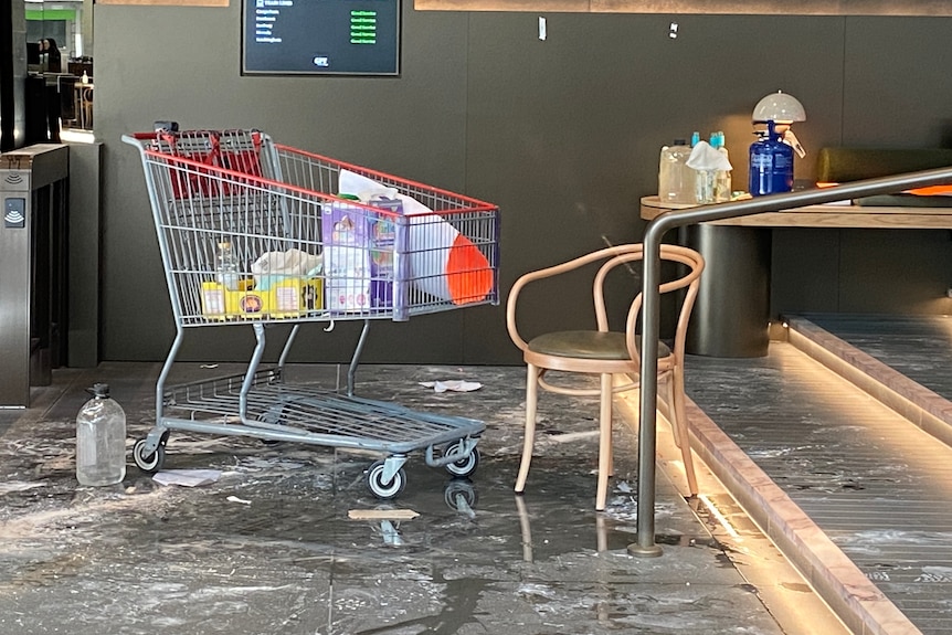 A shopping trolley with a few items inside. There is fluid on the floor and bottles of fluid on a nearby table