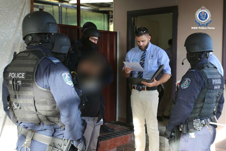 Several police in riot gear surround a man, blurred out, while a detective appears to read him his rights.