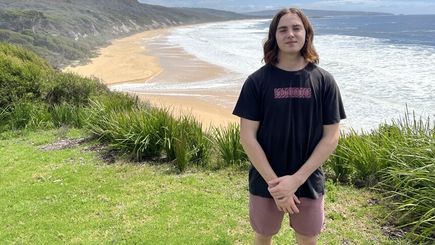 A young man stands on a grassy hill with a beach in the background.