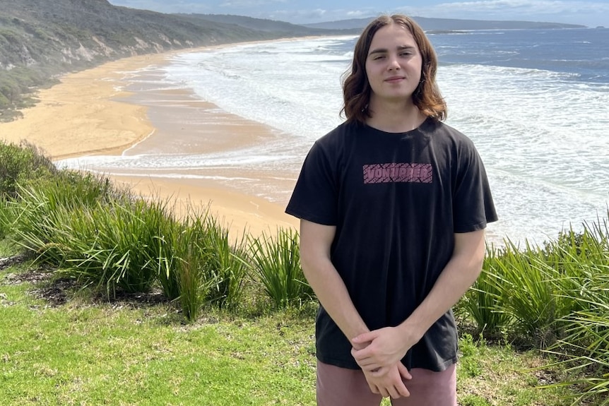 A young man stands on a grassy hill with a beach in the background.