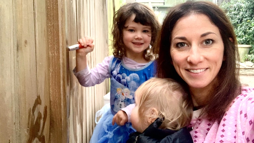 A woman holds a baby while a young child paints a fence with a brush dipped in water. 