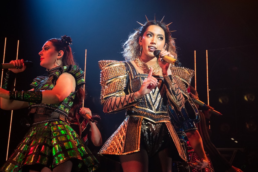 Young woman wearing gold-spiked headband and black mini-dress covered in gold studs with massive shoulders, holding mic, singing