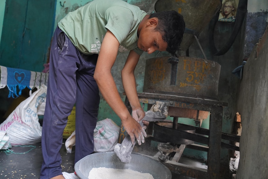 A teenage boy in trackies and a t-shirt grinds flour in a machine 