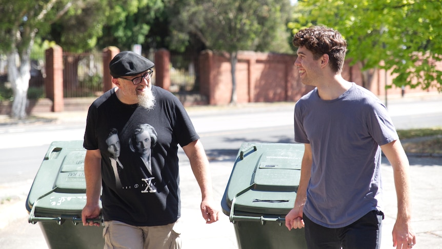 Two men smile at each other as they take wheelie bins in off the side of the road.