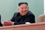 Kim Jong-un smiles as he sits at a long table while others applaud him.
