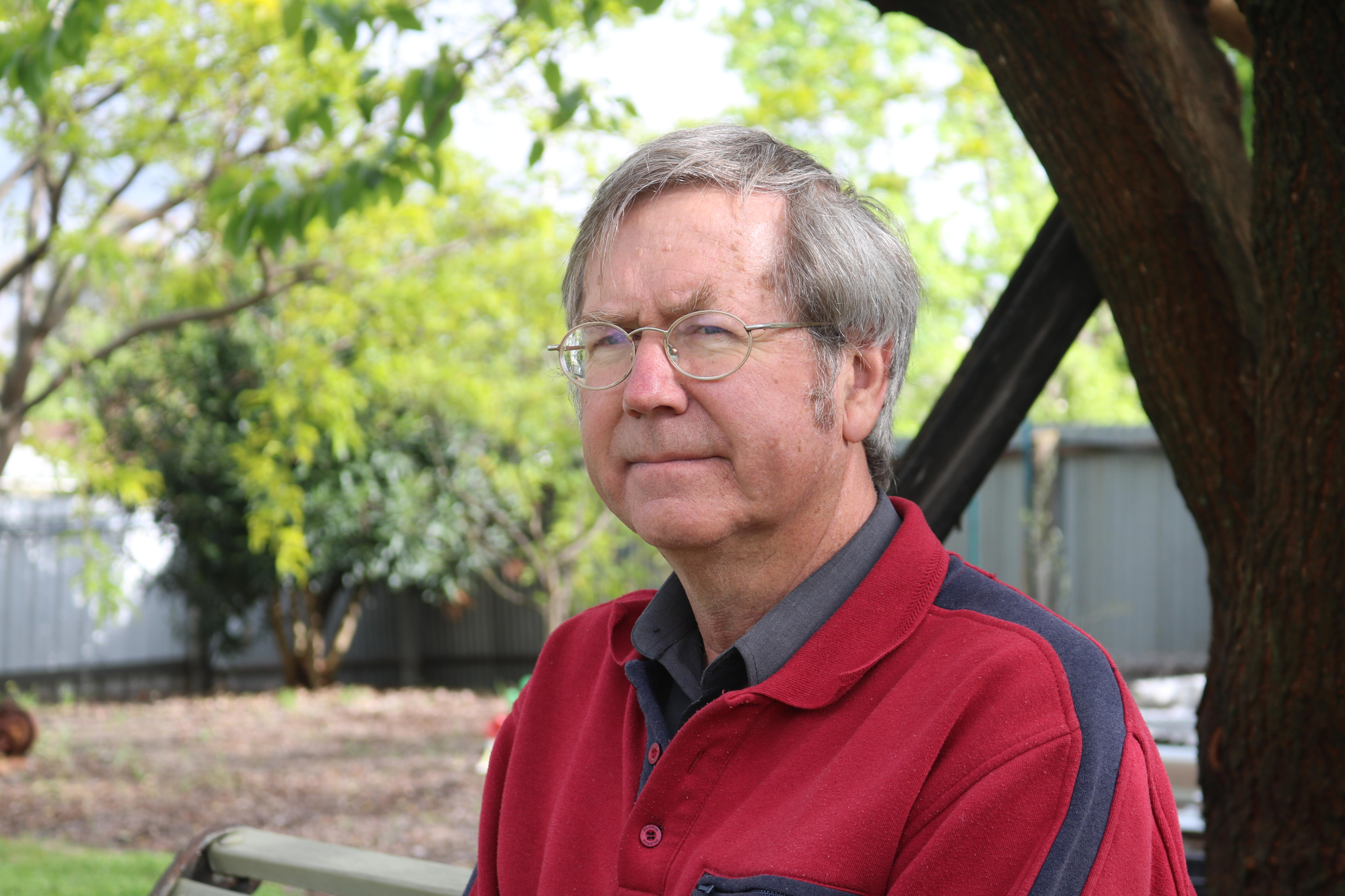 A man with glasses and short hair looks into the distance while sitting on a bench outside under a tree.