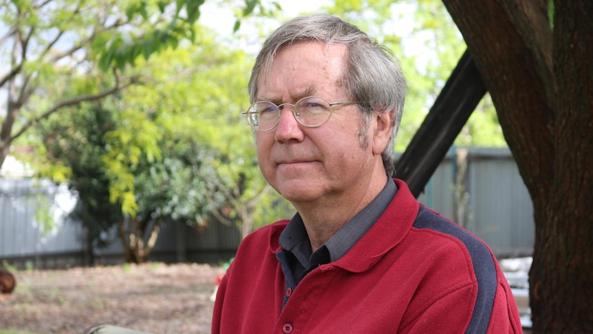 A man with glasses and short hair looks into the distance while sitting on a bench outside under a tree.
