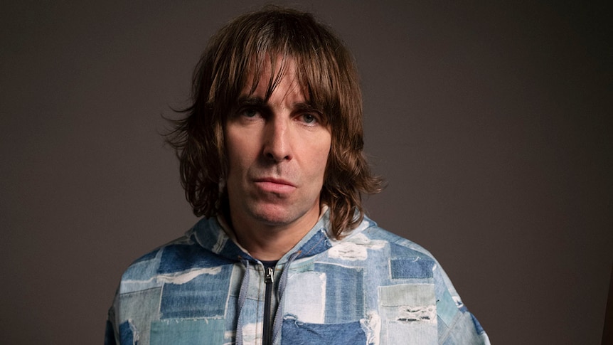 Liam Gallagher with shoulder length hair wears a hoodie with blue squares on it.