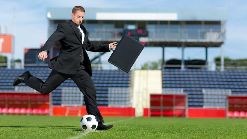 A man in a business suit with briefcase kicks a soccer ball in an empty stadium
